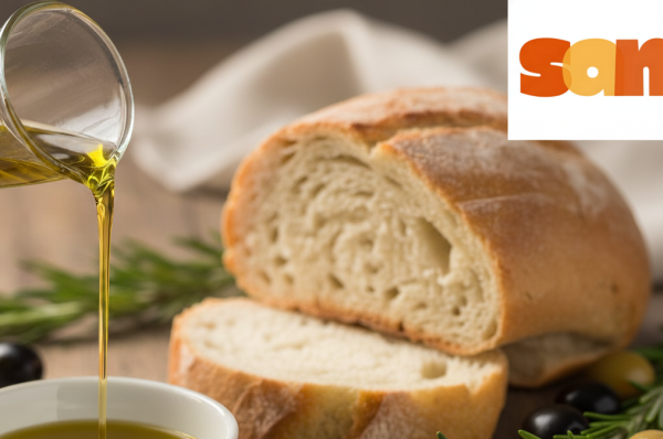 A close-up shot of rich, golden-green Extra Virgin Olive Oil being poured from a glass cruet into a small white dipping bowl. The bowl sits on a rustic wooden table, surrounded by slices of crusty bread, fresh rosemary sprigs, and whole black and green olives. The scene is lit with warm, natural light.