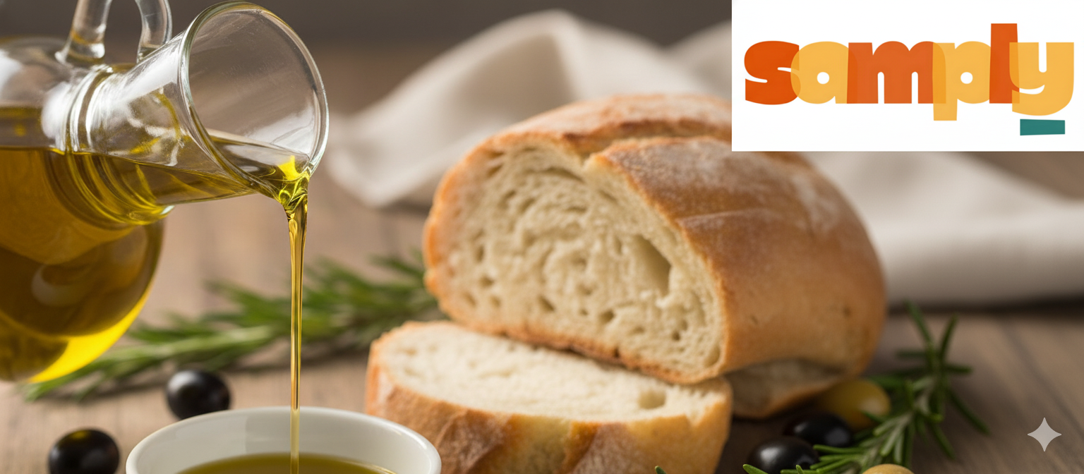 A close-up shot of rich, golden-green Extra Virgin Olive Oil being poured from a glass cruet into a small white dipping bowl. The bowl sits on a rustic wooden table, surrounded by slices of crusty bread, fresh rosemary sprigs, and whole black and green olives. The scene is lit with warm, natural light.
